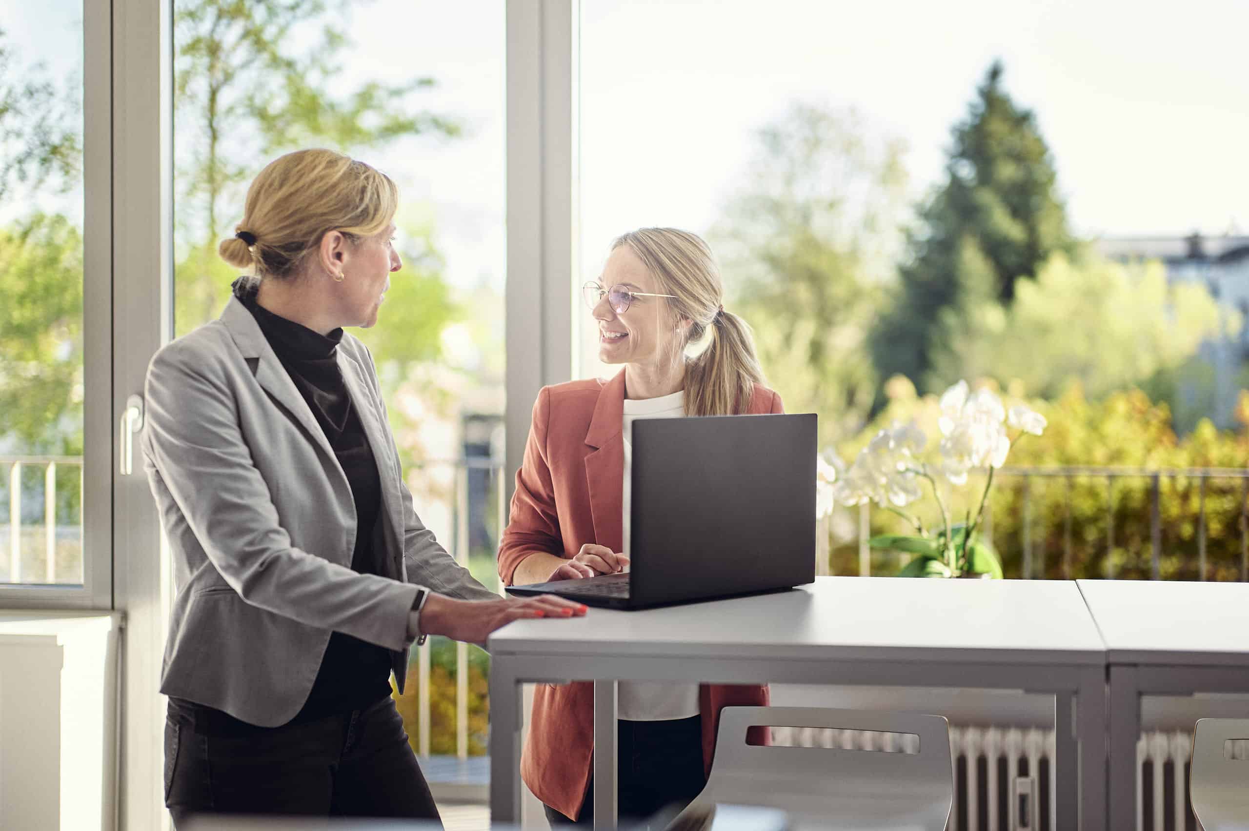 Celine Spitz und Magdalena Sabaß stehen in einer Lounge an einem Bartisch, auf dem ein Laptop liegt, und unterhalten sich miteinander.