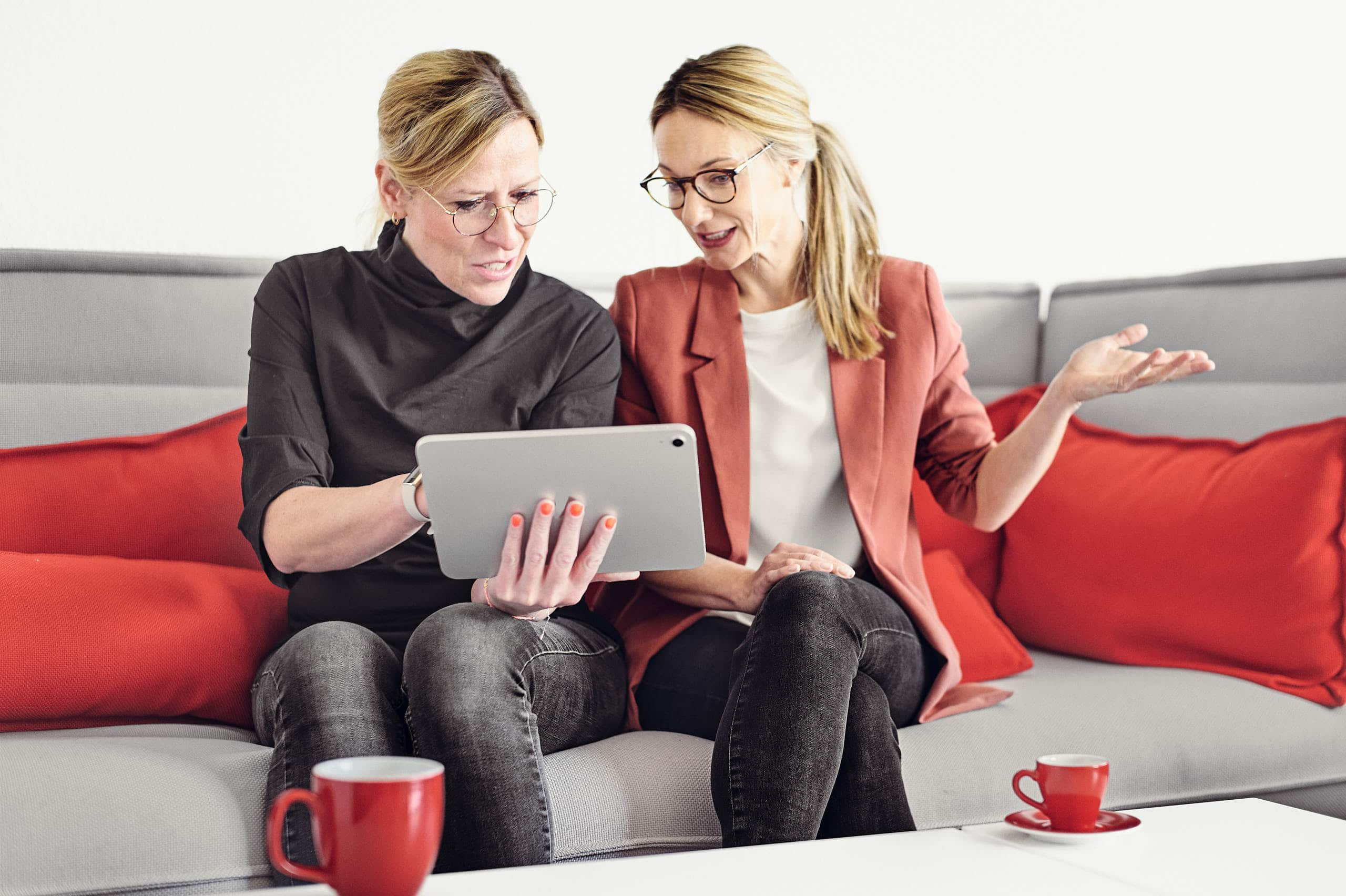 Magdalena Sabaß erklärt Celine Spitz etwas auf einem Tablet, während die beiden auf einem Sofa sitzen und auf dem Couchtisch steht eine Tasse Kaffee.