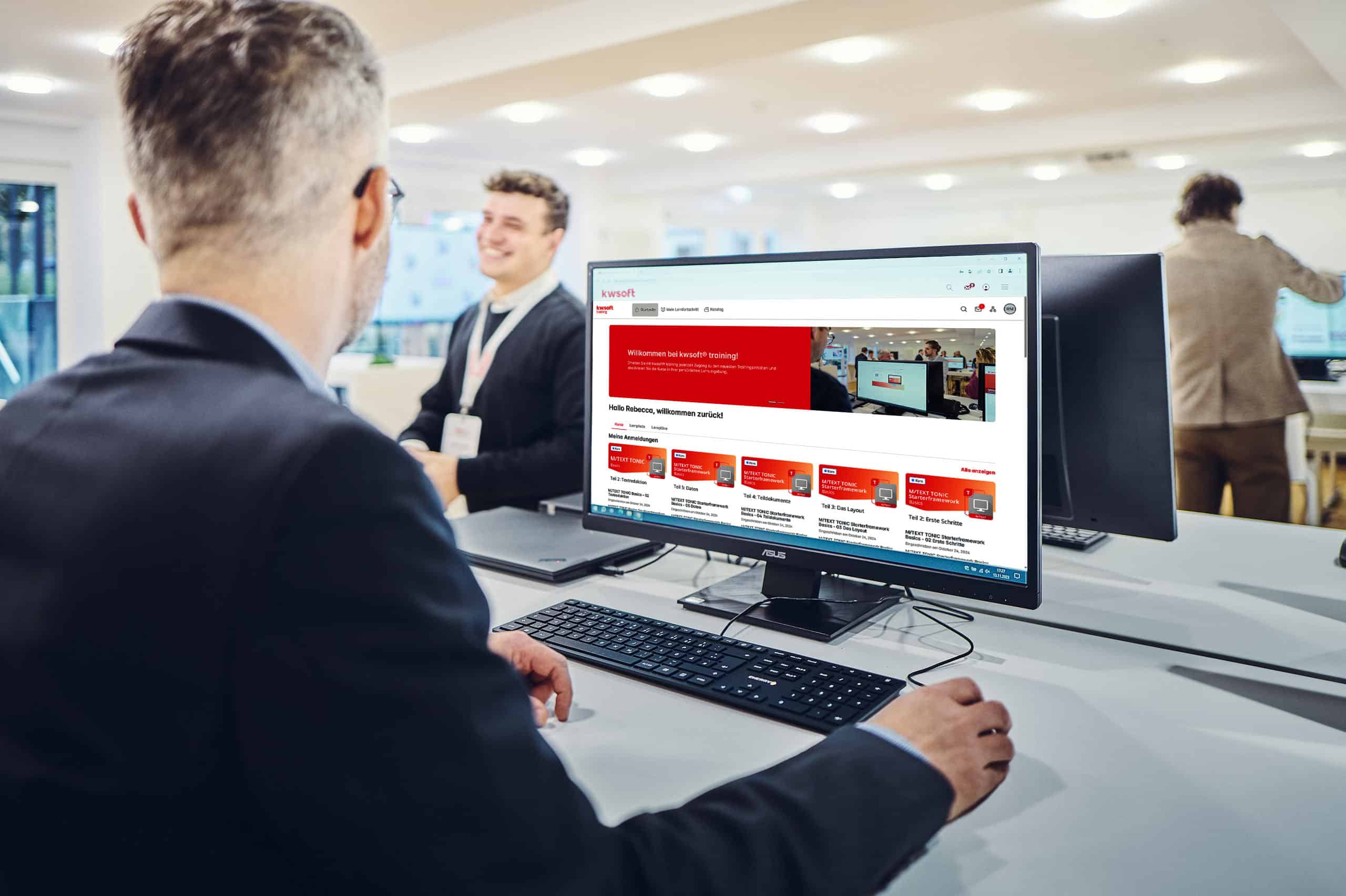 Ein Mann sitzt vor einem Bildschirm, auf dem eine kwsoft-Schulung läuft, und schaut zu einem Mann, der im Hintergrund steht.